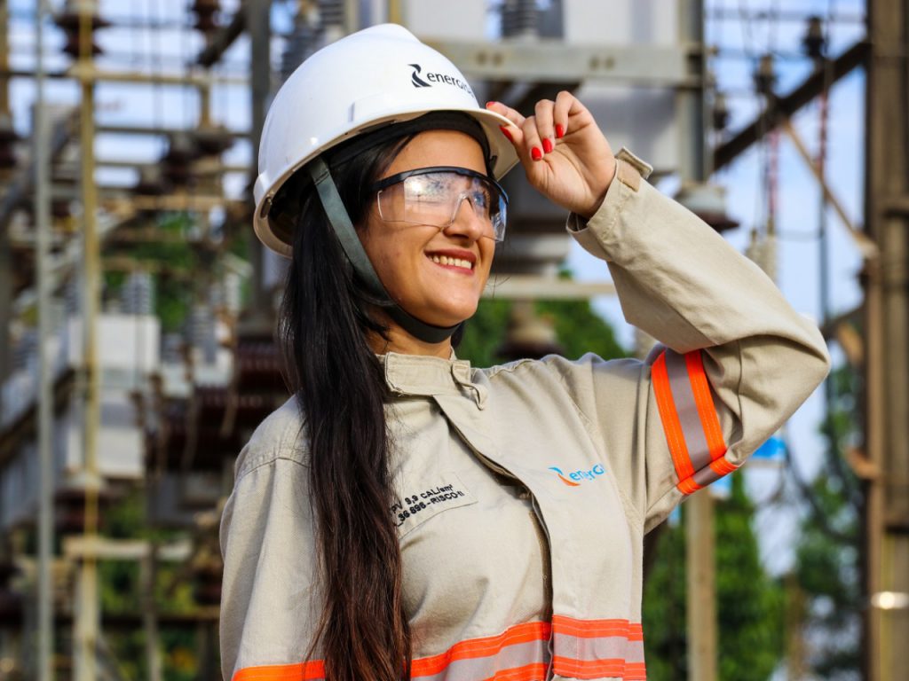 Colaboradora sorri para o horizonte, com mão segurança aba do capacete. 